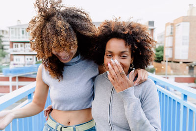 Positive african american female friends with curly hair embracing and laughing in city street while enjoying weekend and having fun