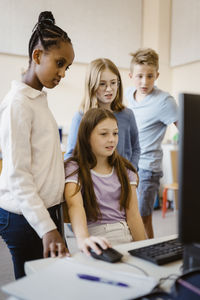 Girl using computer while sitting amidst male and female pupils in classroom