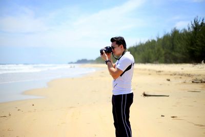 Man photographing through camera while standing on sand at beach