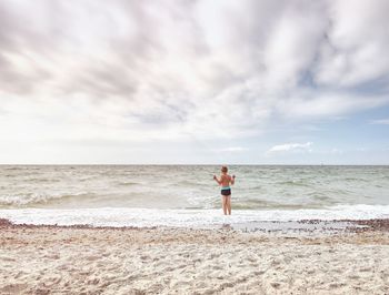 Blond hair boy stay in cold sea tide. kid on stony beach with foamy waves. windy day, cloudy sky