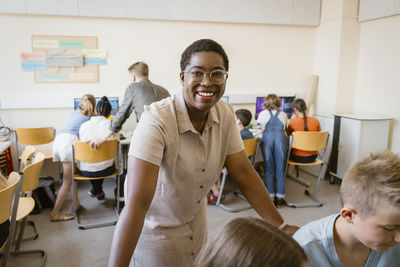 Portrait of female teacher leaning on chairs with students in computer class at school