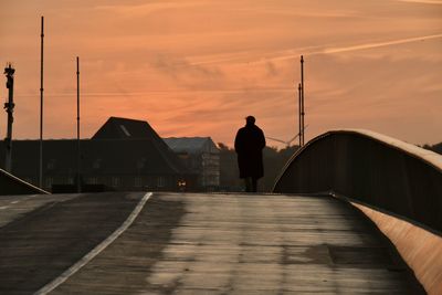 Rear view of man standing by railing against sky during sunset