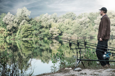 Side view of man looking at lake while standing by fishing rod 