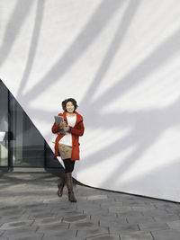 Curly woman in red coat is walking by white wall. student with laptop. freelancer with device.