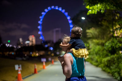 Mother carrying daughter on shoulder on footpath in city