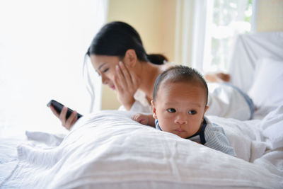 Cute baby lying by mother using phone on bed 