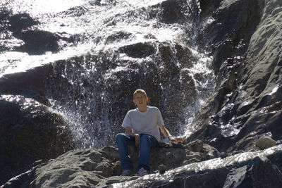 Low angle view of teenage boy sitting on rock formation against waterfall at banff national park