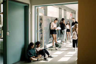 Girl and boy sitting in doorway while professor talking to students in school corridor