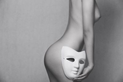 Midsection of naked woman holding mask while standing over gray background