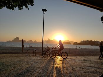 Silhouette people riding bicycle on beach against sky during sunset