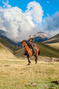 Side view of a cowgirl shepherd riding on a horse on a mountain valley against sky with clouds