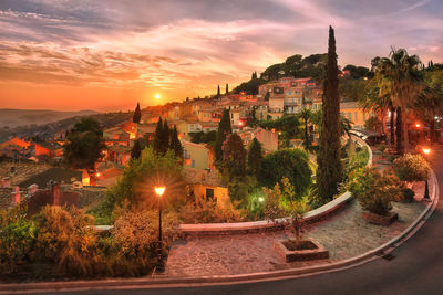 Trees and houses in illuminated town against sky during sunset