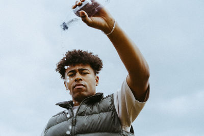 Low angle portrait of young man pouring water from bottle against sky