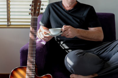 Midsection of man holding coffee while sitting by guitar at home
