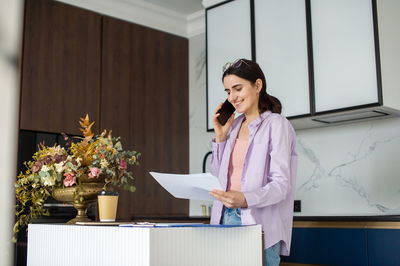 A cute girl in a shirt is standing near the table, holding sheets of paper and talking on the phone
