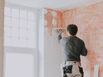 A young caucasian guy applies fresh putty with a small spatula on the wall near the window