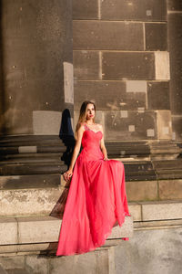 Full length of young woman in pink evening gown sitting against columns