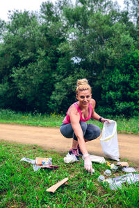 Full length portrait of woman picking up garbage on grass against trees