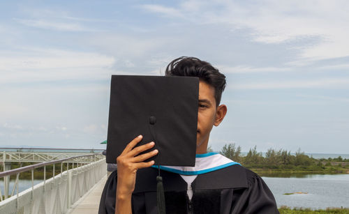 Portrait of young man holding graduation gown by lake against sky