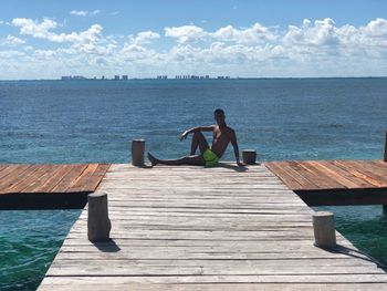 Shirtless man sitting on pier at sea against sky