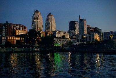 Illuminated buildings by river against sky in city