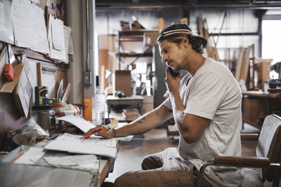 Side view of carpenter using mobile phone while reading document in workshop