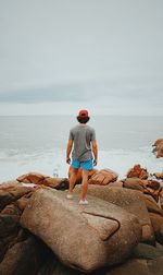 Rear view of mid adult man standing on rock at beach against cloudy sky