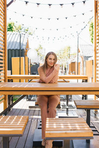 A smiling young attractive woman poses sitting at a table of an outdoor cafe on a summer terrace