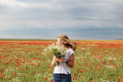 Young woman holding flowers while standing on field against cloudy sky