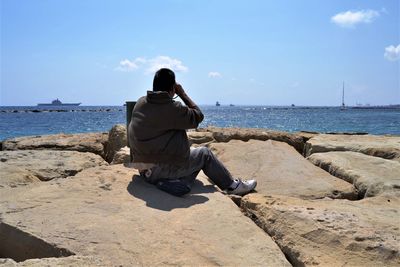 Full length of man looking at sea through binoculars on rocks against sky