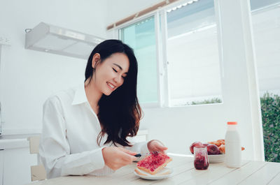 Low angle view of happy woman applying preserves on bread slice at table during morning