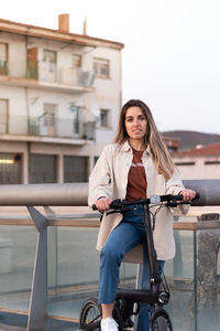 Vertical portrait of young woman looking at camera holding an electric bicycle in urban enviroment