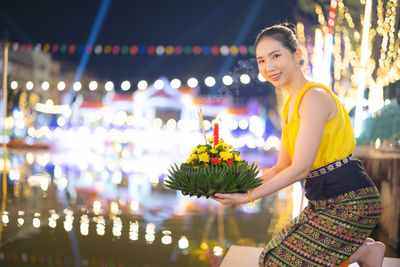 Portrait of young woman standing by illuminated christmas tree at night