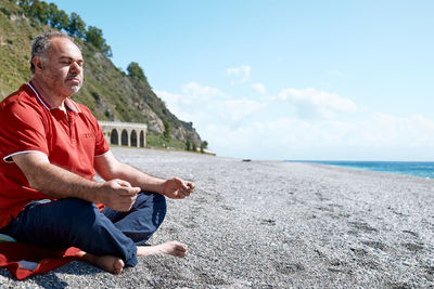 Full length of bearded man in casual clothes sitting in lotus pose on pebble beach and meditating