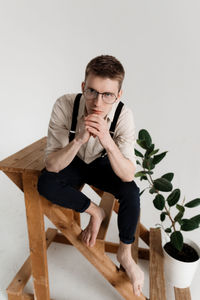 Portrait of young man sitting by potted plant on ladder against white background