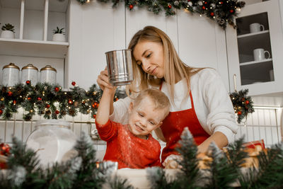 Young smiling mother cooking with little son and sifting flour with sieve in the decorated kitchen