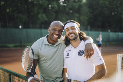 Happy man with arm around male friend wearing headband at tennis court