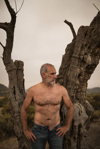 Shirtless adult man standing by bare olive tree trunk against clear sky 