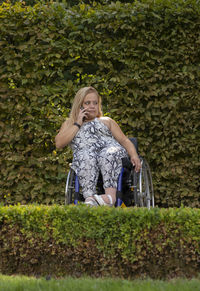 Full length young woman on wheelchair talks on phone in green park at summer day. busy female adult