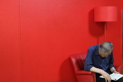 Senior man reading book while sitting on chair by red wall at home