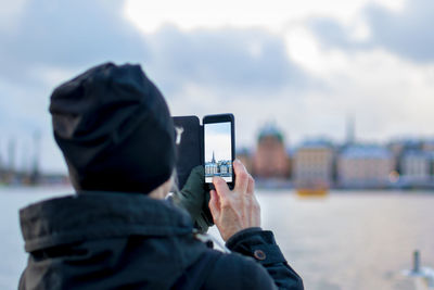 Rear view of man photographing buildings from mobile phone against sky