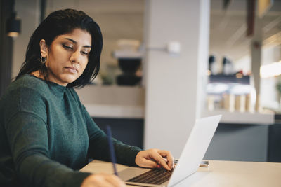 Businesswoman writing while using laptop at desk in office