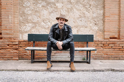 Portrait of young man sitting on bench against brick wall