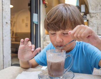 A ten-year-old caucasian boy is sitting in a street cafe and drinking cocoa with a teaspoon.