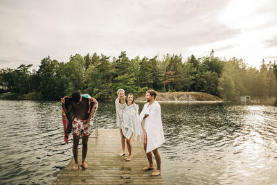 Happy friends wearing towel while standing on jetty at lake during vacation
