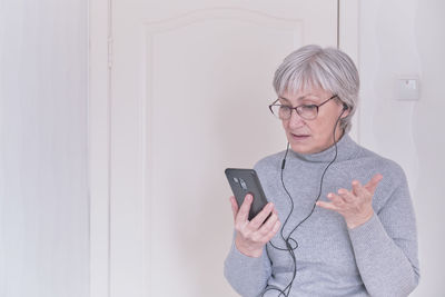 A gray-haired senior woman with glasses and a gray turtleneck talking on mobile phone at home. 