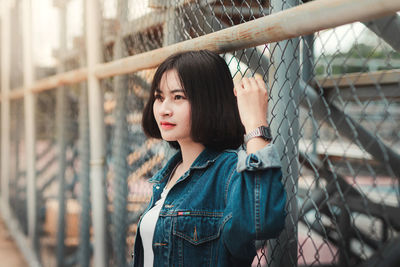 Beautiful young woman looking away while standing by chainlink fence