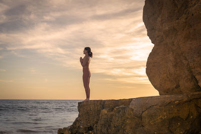 Woman standing on rocks by the sea practicing yoga at sunrise or sunset.