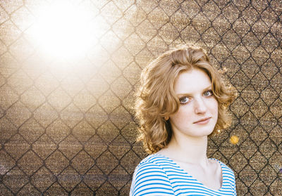 Portrait of young woman standing by chainlink fence against fabric