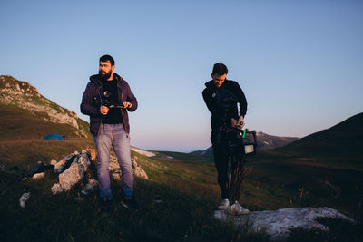 Full length of men holding camera while standing on mountain against clear sky